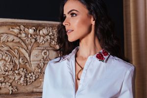 Woman with dark hair wearing a crisp white shirt with floral embroidery detail poses against an ornate carved wall.