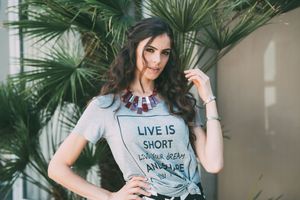 A person wearing a gray t-shirt with 'Life is Short' text poses against a backdrop of palm leaves.