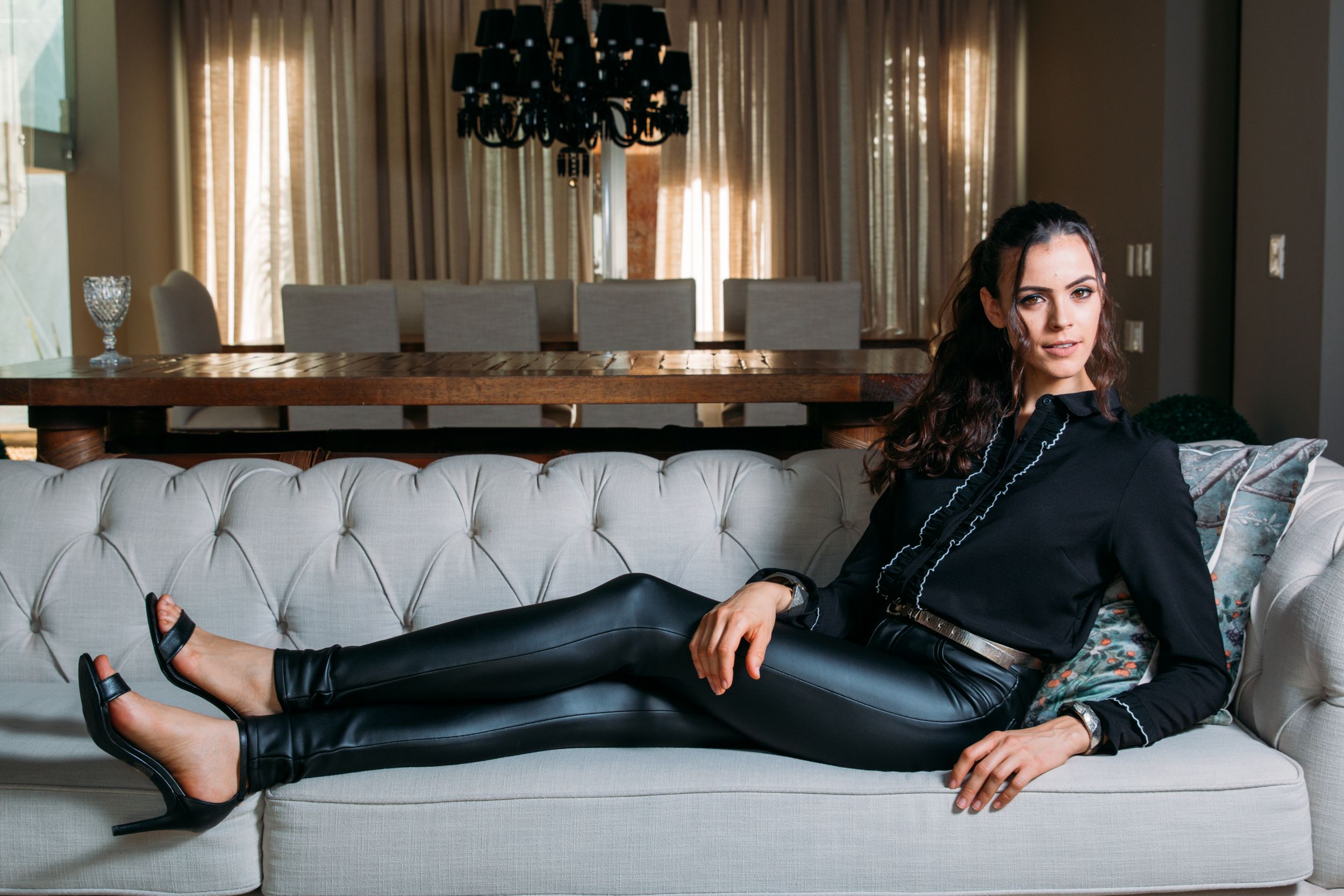 A person in a black leather outfit reclines on a gray tufted sofa in an elegant living room setting.