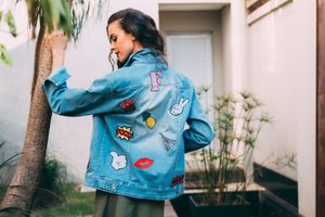 Person wearing a light blue denim jacket decorated with colorful patches and pins stands near a tree.