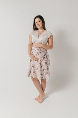 A pregnant woman in a flowy floral dress poses barefoot against a white background during a maternity photo shoot.