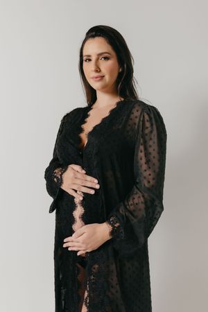Woman wearing black lace maternity robe stands against white backdrop in elegant pregnancy photoshoot.