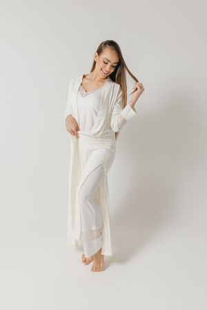 Person in white loungewear poses wearing long robe and pajama set against minimal background while playing with hair.
