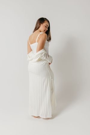 A model showcases a flowing white pleated dress with thin shoulder straps in a minimalist studio setting against a white backdrop.
