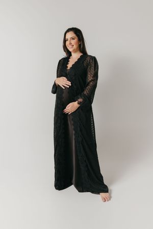 A barefoot pregnant woman poses in a flowing black lace maternity robe against a light gray studio backdrop.
