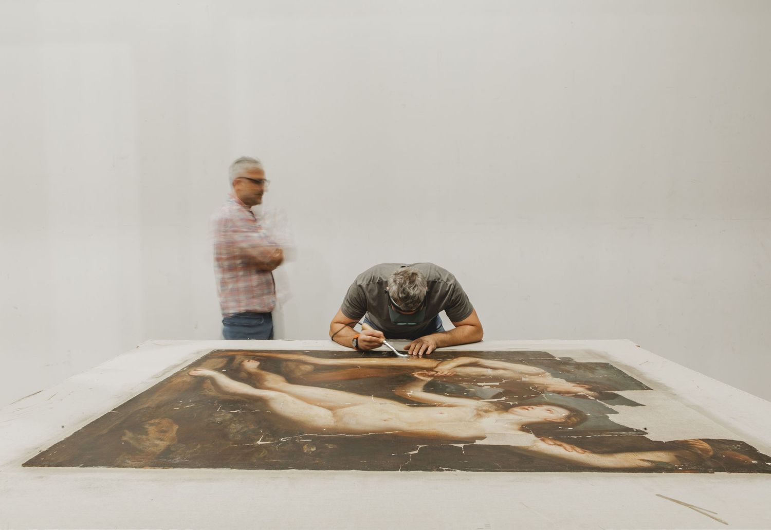 Two artists examine a large art piece laid out on the floor in a bright white studio space.