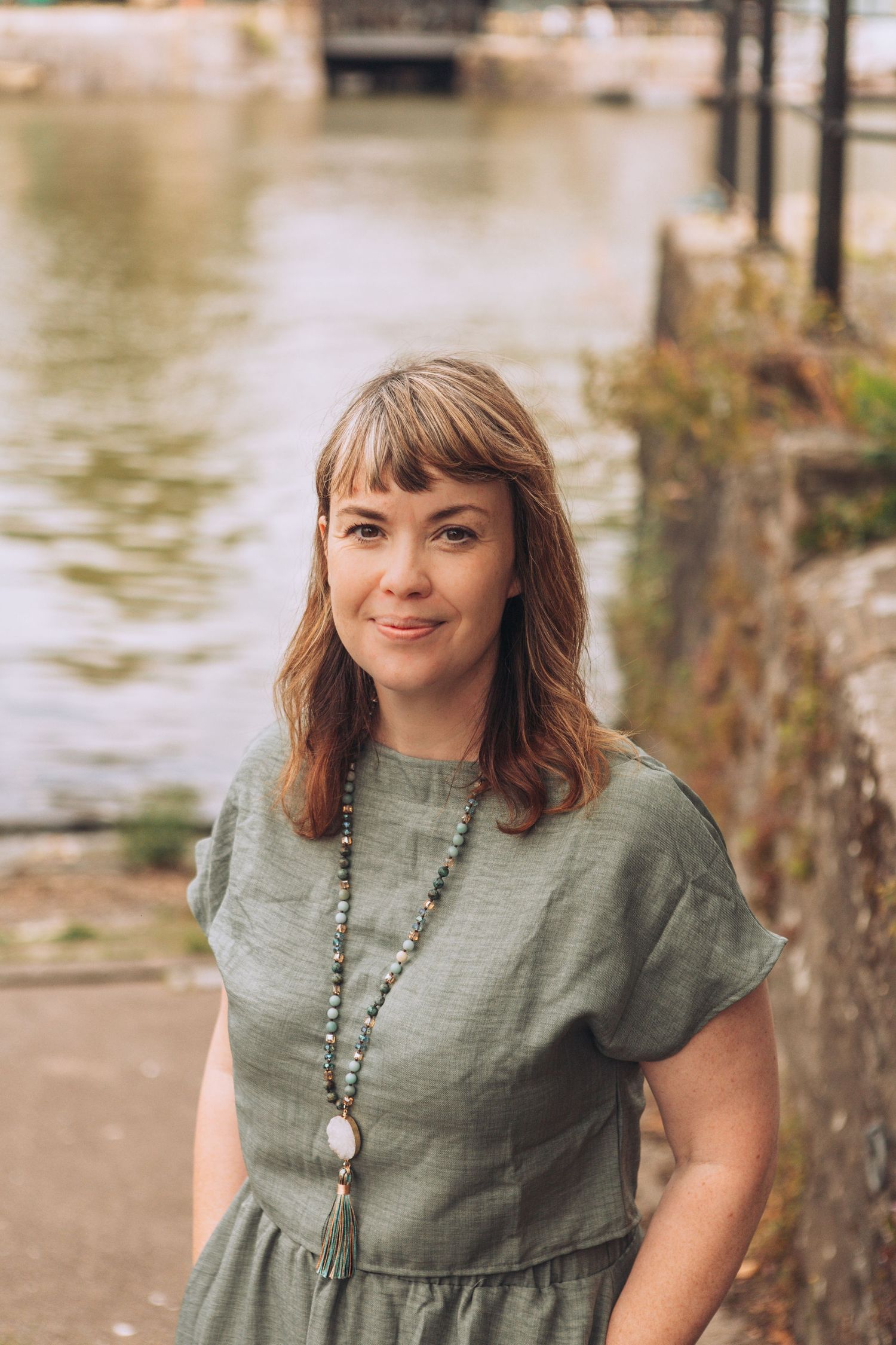 A person in a sage green dress poses outdoors near water on a sunny day.
