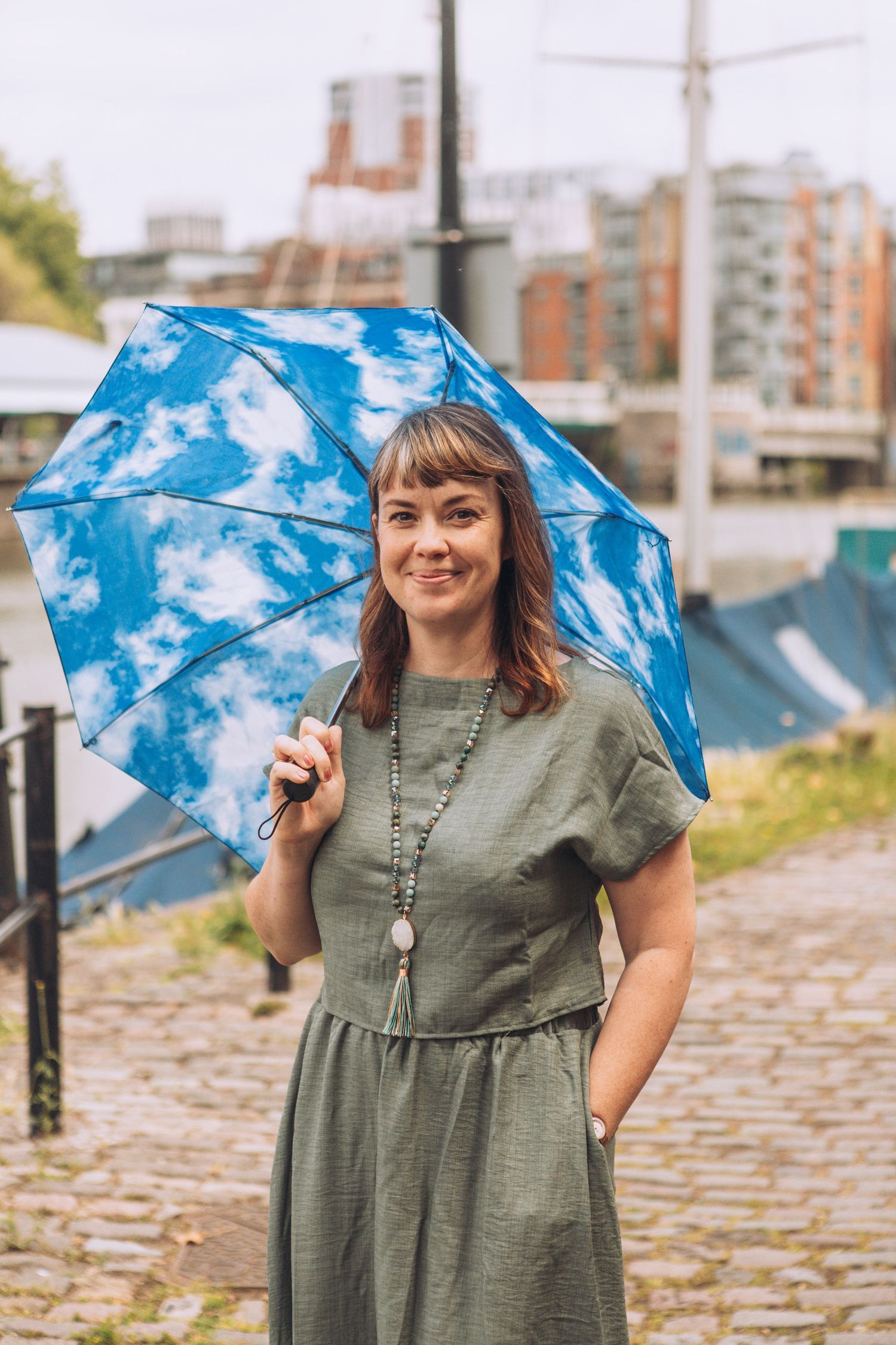 A woman in a green dress poses playfully with a blue umbrella on a cobblestone street.