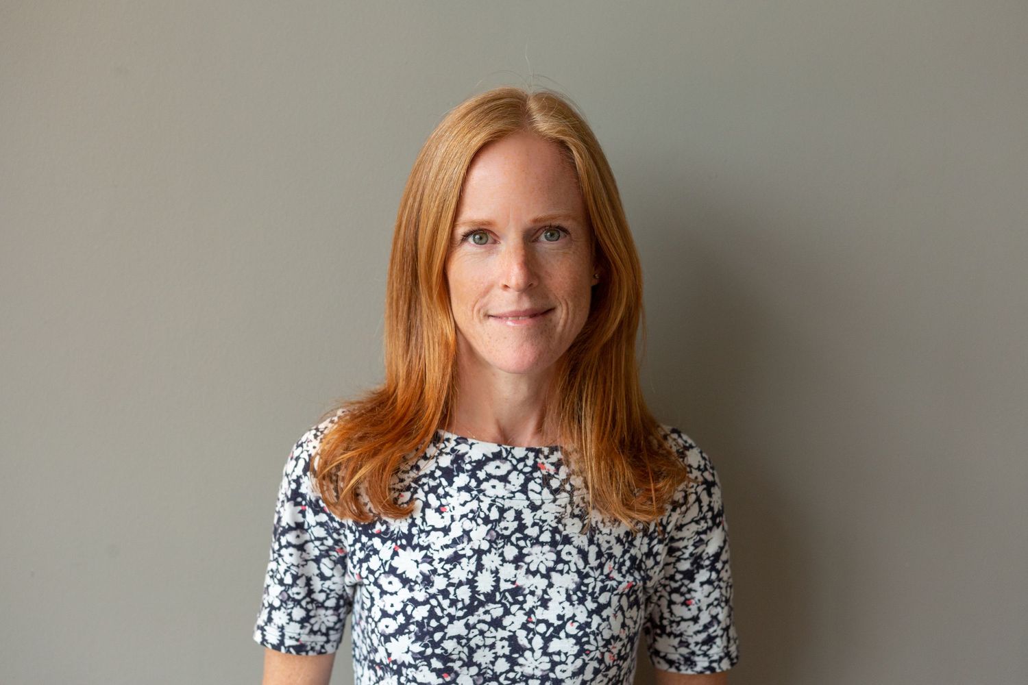 Woman with red hair wearing a floral black and white dress stands against neutral wall in portrait series.