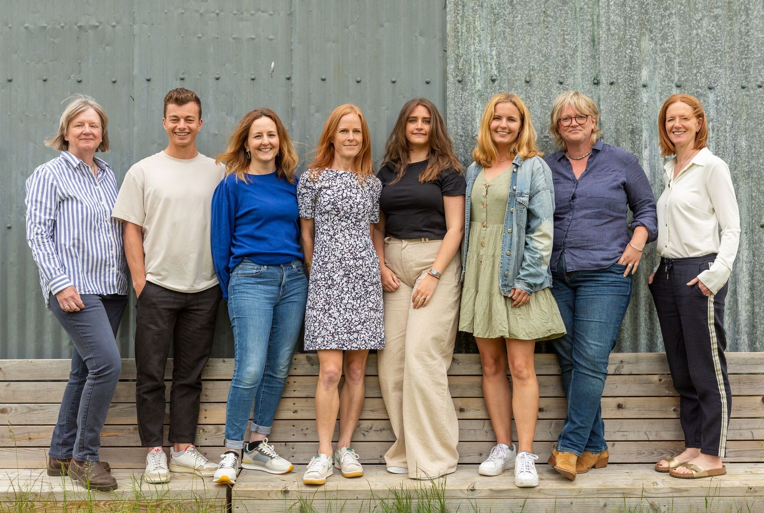 A group of eight people standing together on outdoor wooden steps against a gray wall.