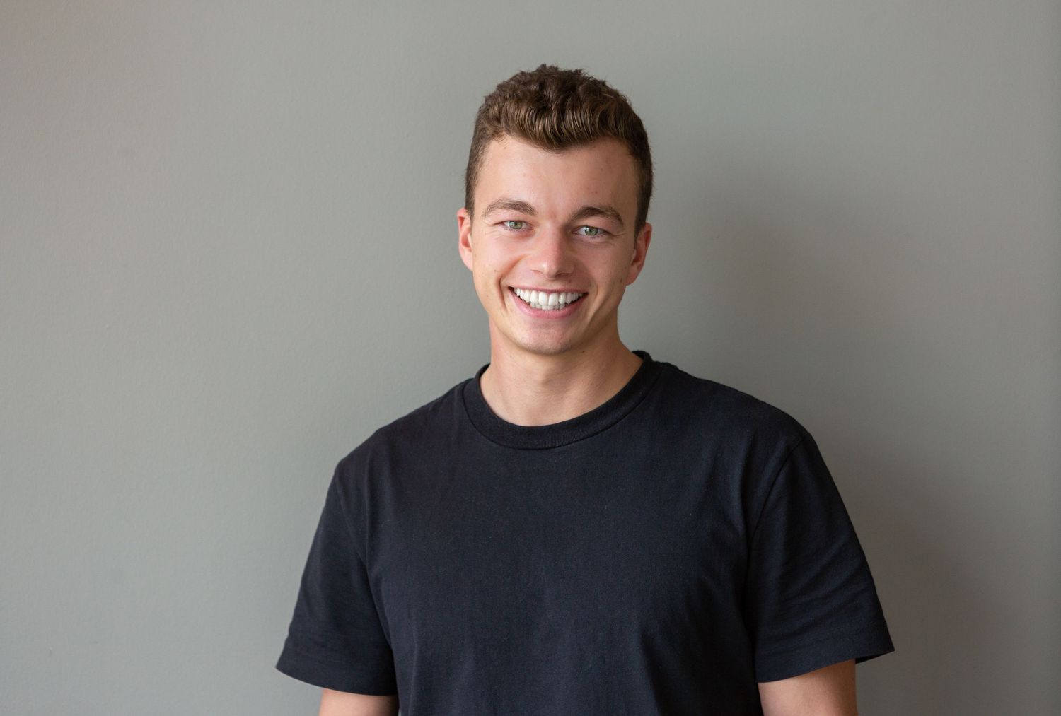 Young adult in black t-shirt smiling confidently against gray wall.