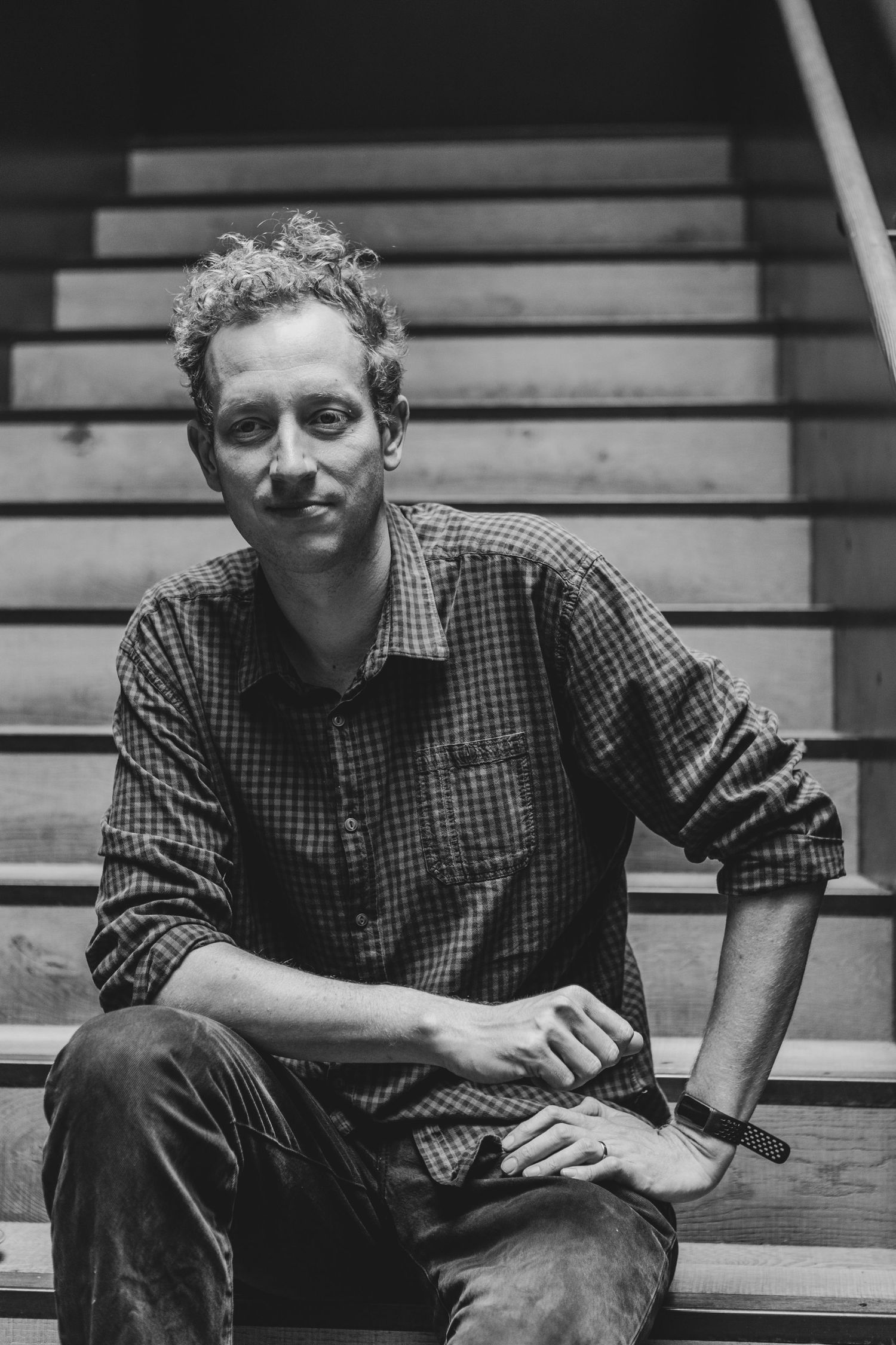 A black and white portrait of someone sitting on wooden stairs wearing a checkered button-down shirt.