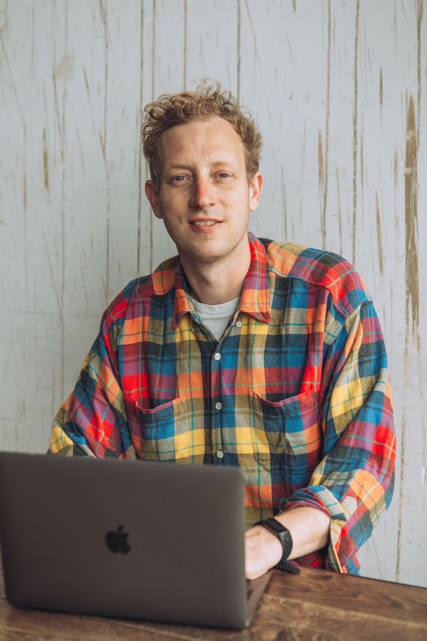 Four progressive photos show someone in a colorful plaid shirt working on a MacBook against a white wooden wall.