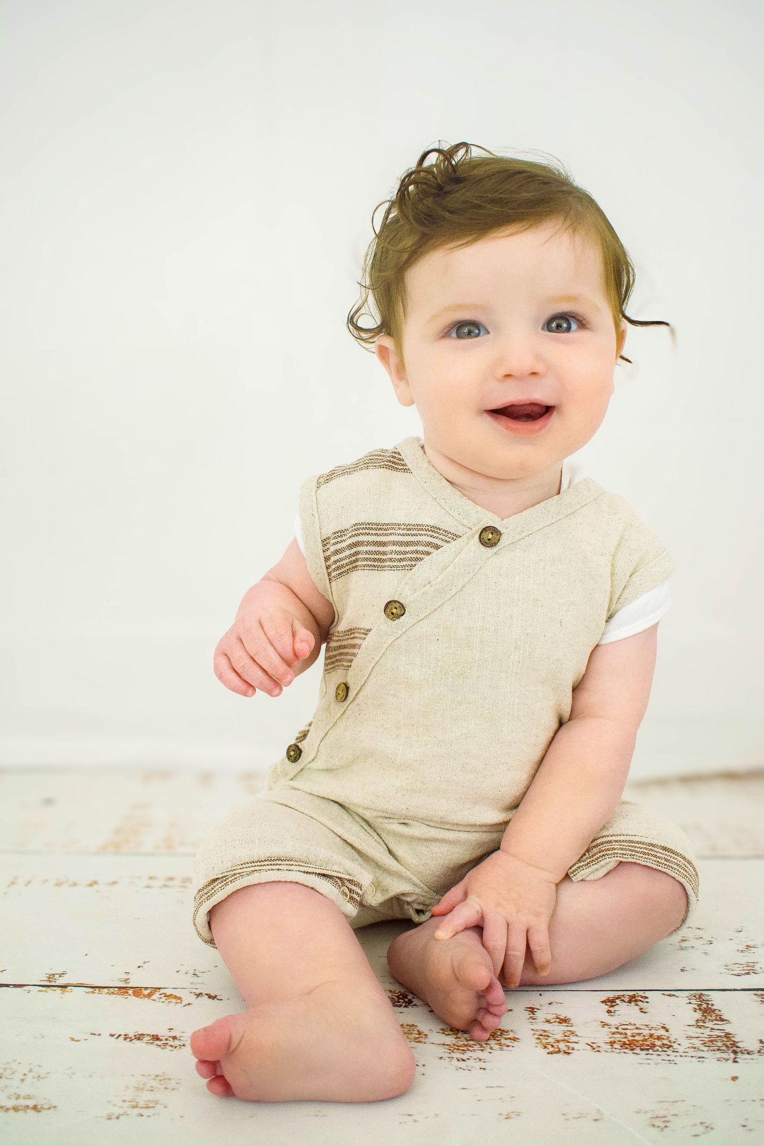 Baby in beige romper sitting on distressed white wooden floor.