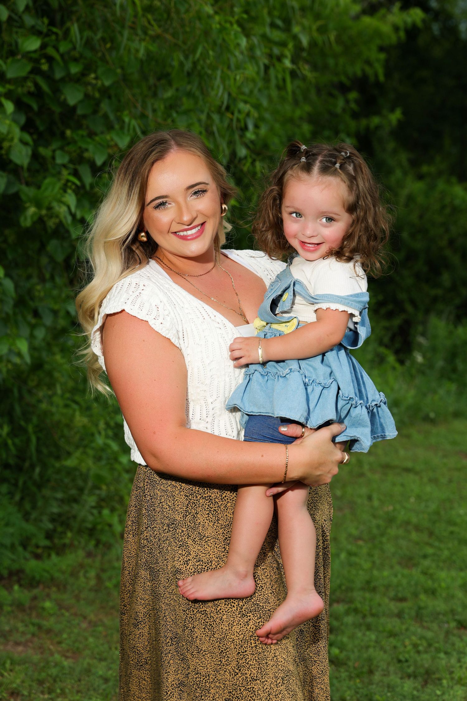 A woman in a white top and brown skirt holds a small child wearing denim against a green outdoor backdrop.