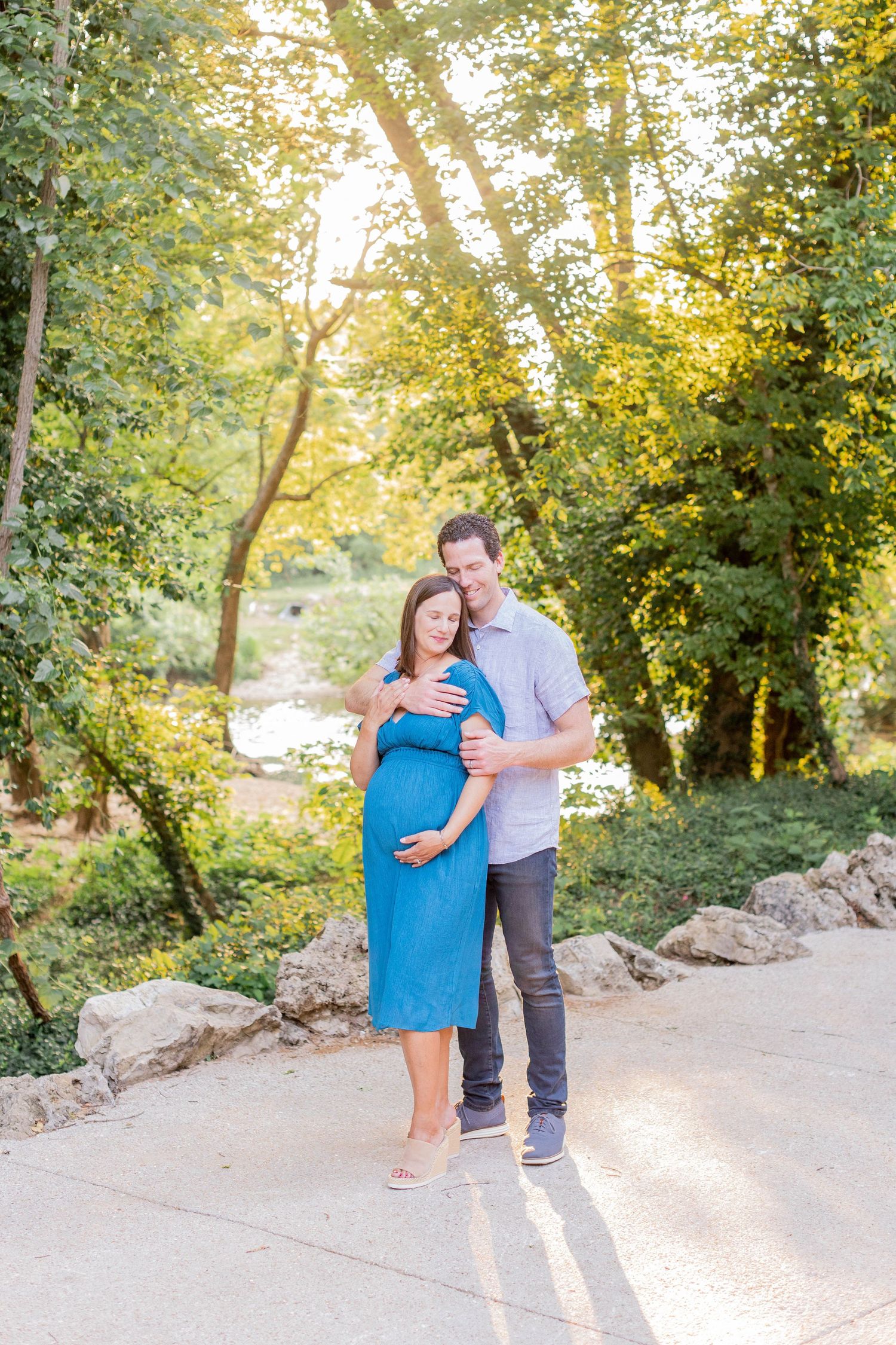 A couple sharing an intimate moment on a sunlit path surrounded by lush trees and greenery on a bright summer day.