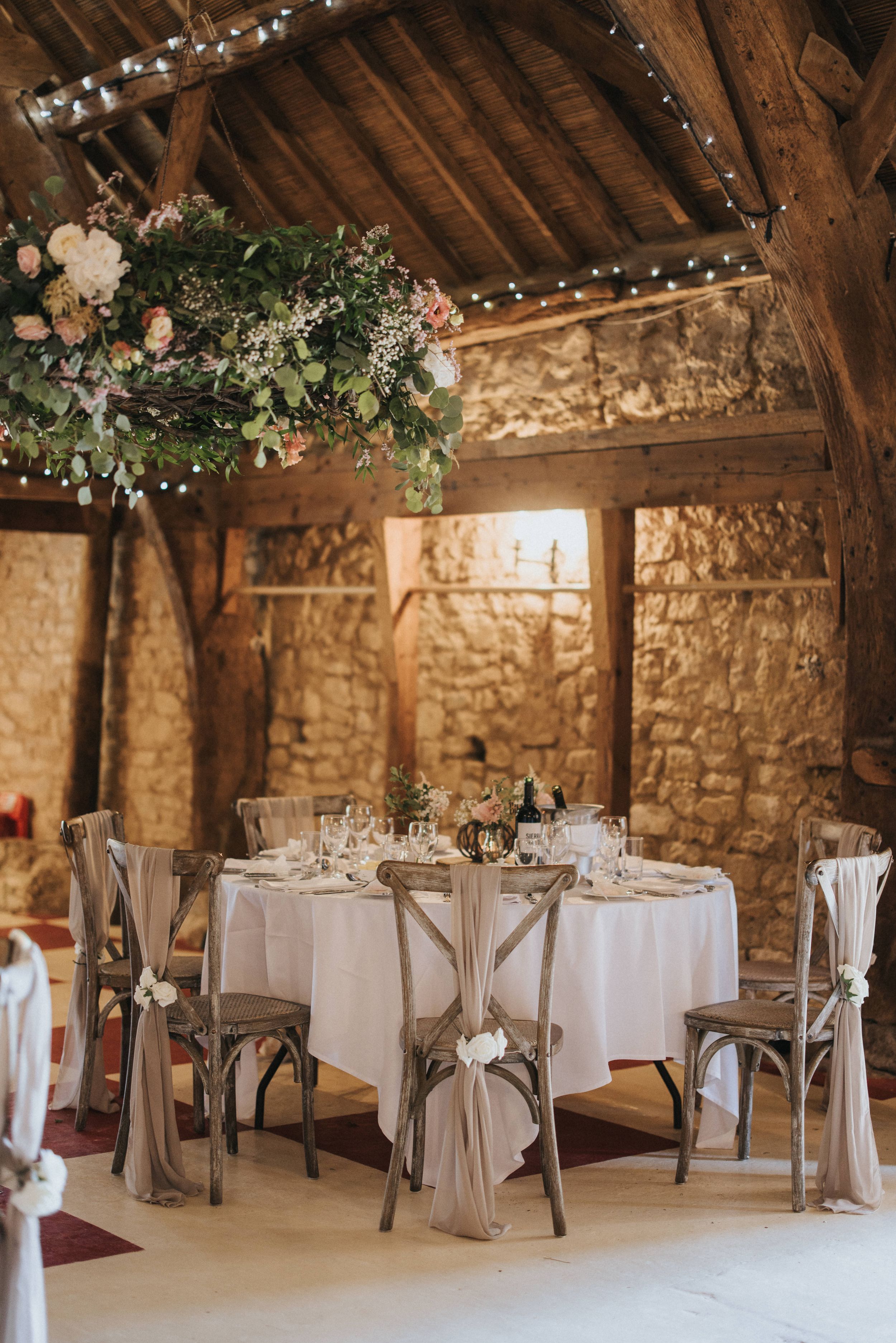 Rustic barn reception venue with white round tables, wooden chairs, stone walls and twinkling lights strung across beamed ceiling.