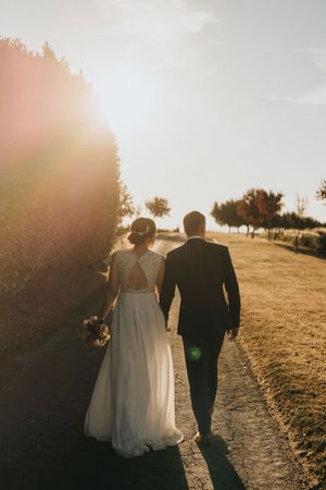 A bride and groom walk along a path at sunset, casting long shadows as sunlight streams through trees.