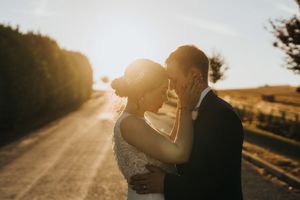 A romantic couple embraces on a sunlit country road during golden hour with lens flare in the background.