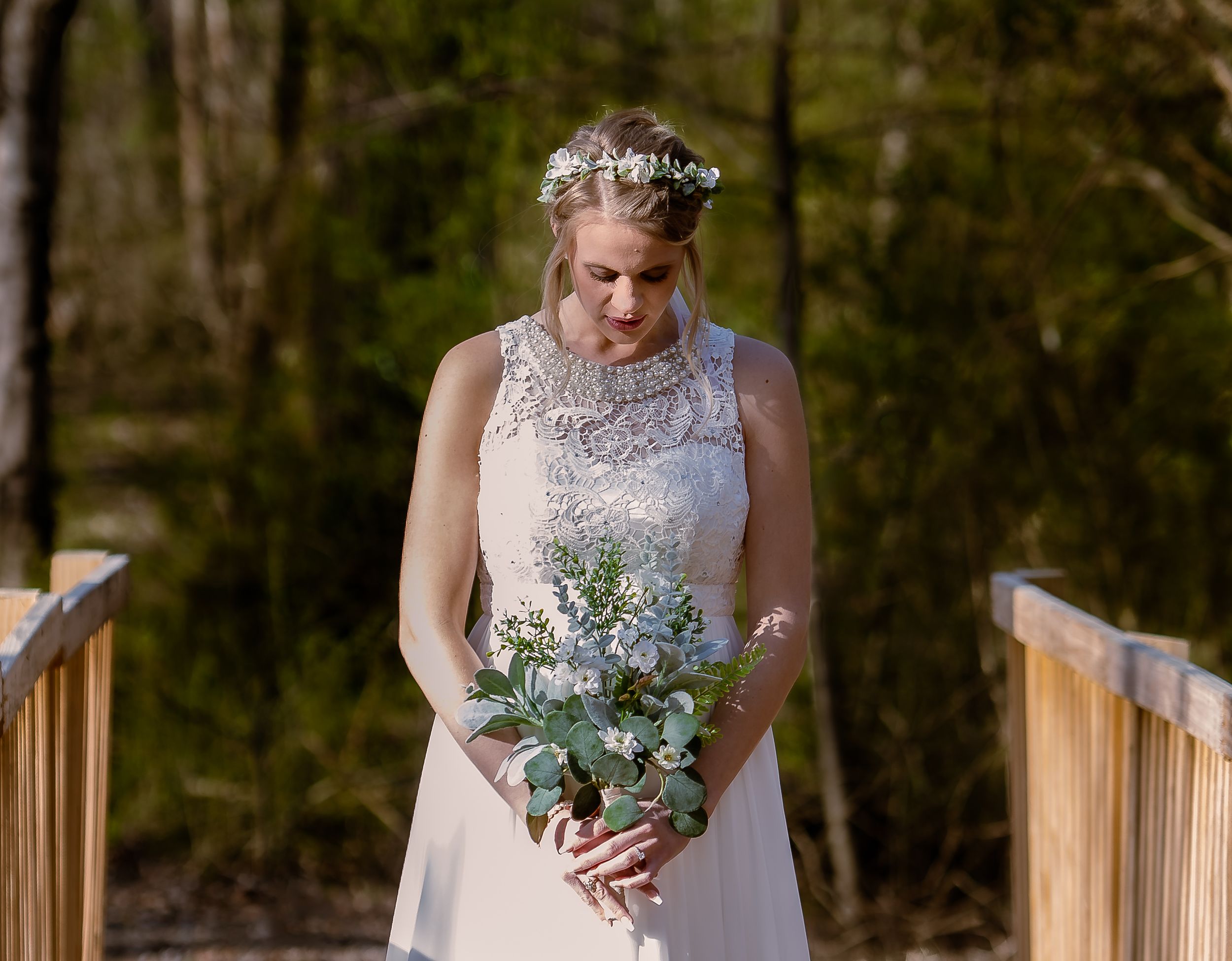 A bride in a lace sleeveless wedding dress looks down at her rustic bouquet of white and green flowers on a wooden deck.