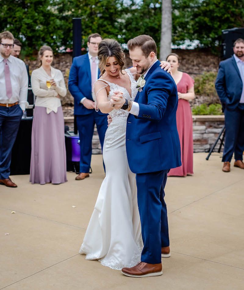 Couple in wedding attire dancing together on an outdoor patio while guests look on in pink and blue attire.
