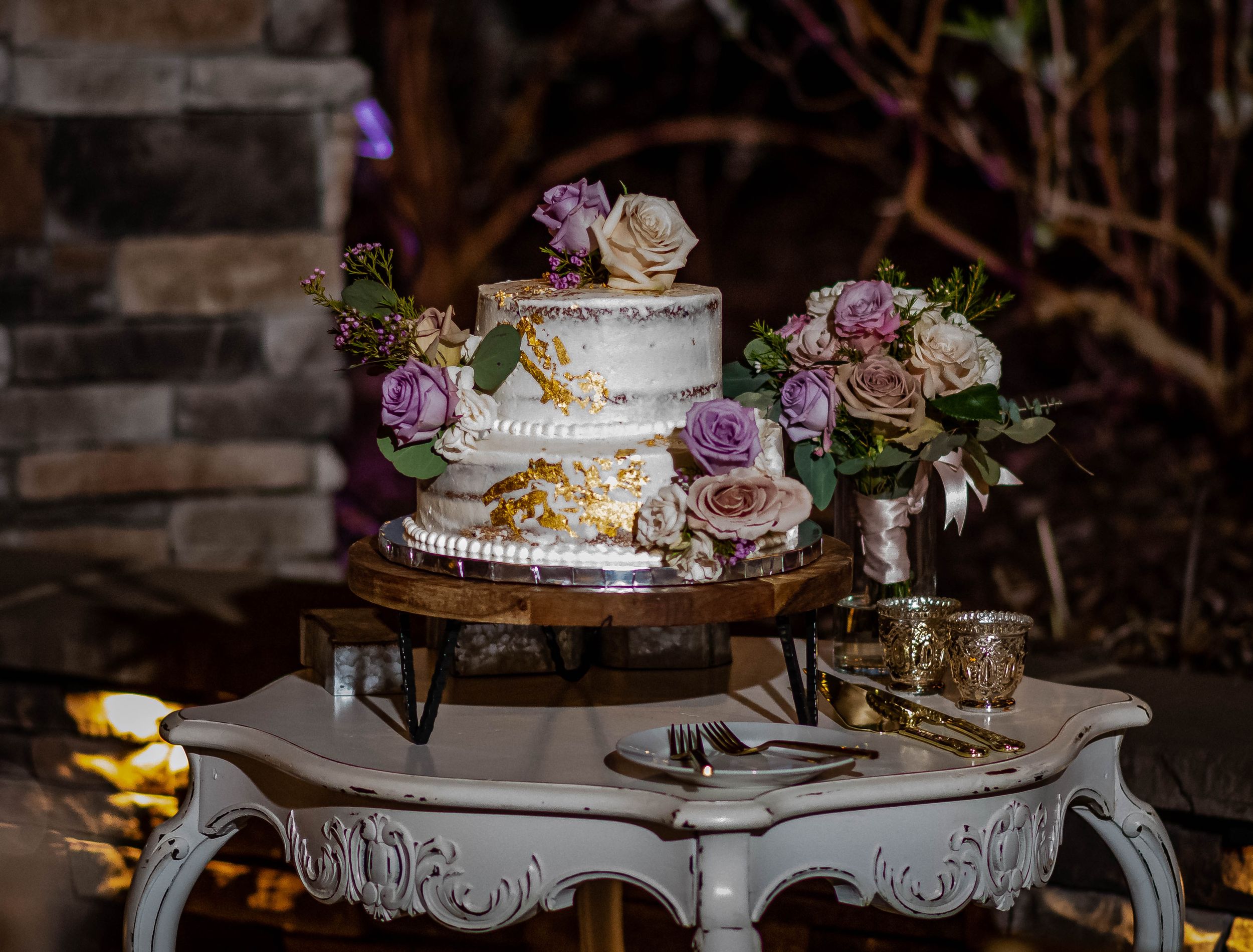 Elegant white wedding cake with mauve roses and gold accents displayed on ornate antique-style pedestal stand.