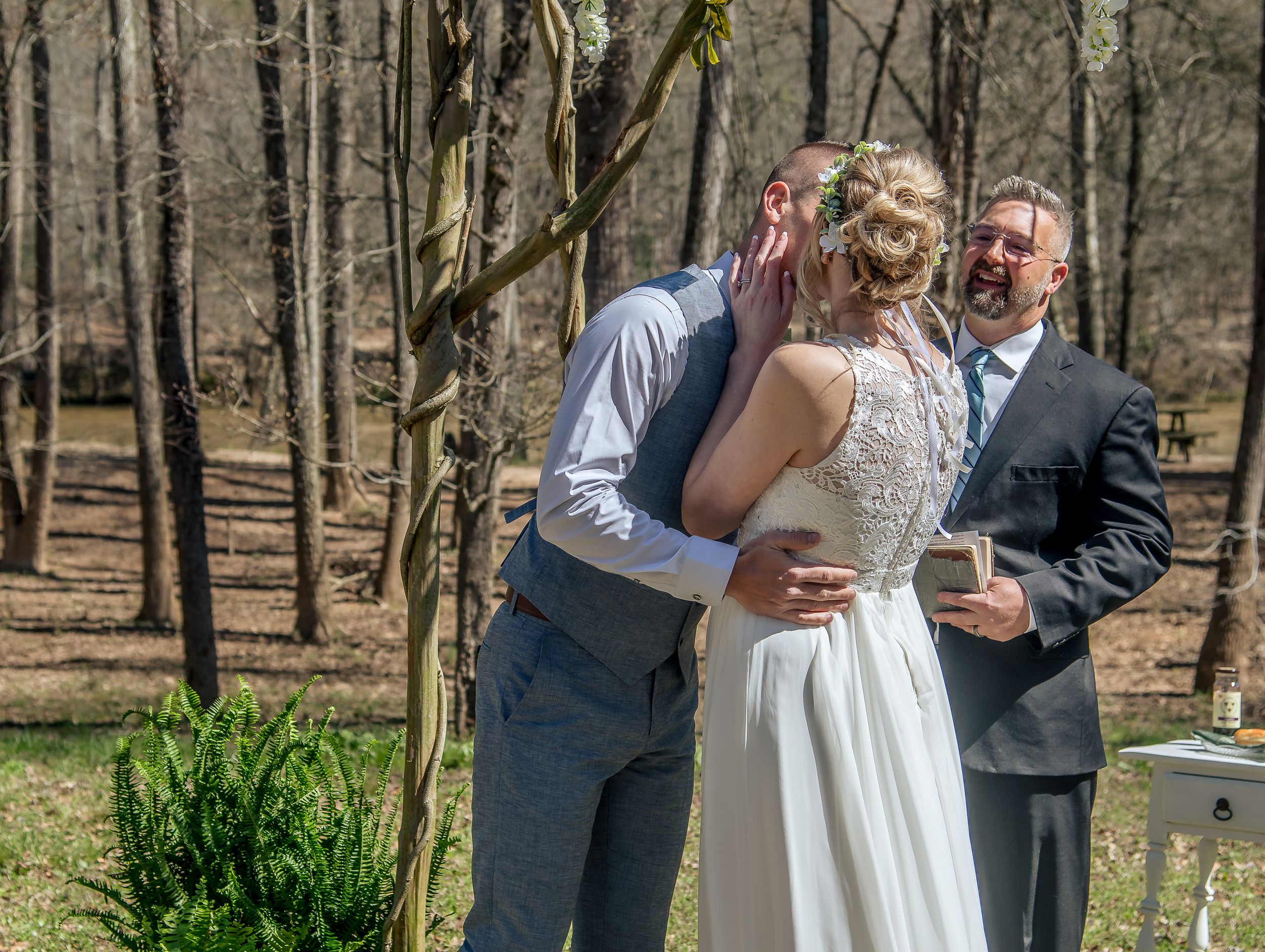 A wedding couple laughs together while dancing in a wooded outdoor setting, surrounded by trees and natural light.