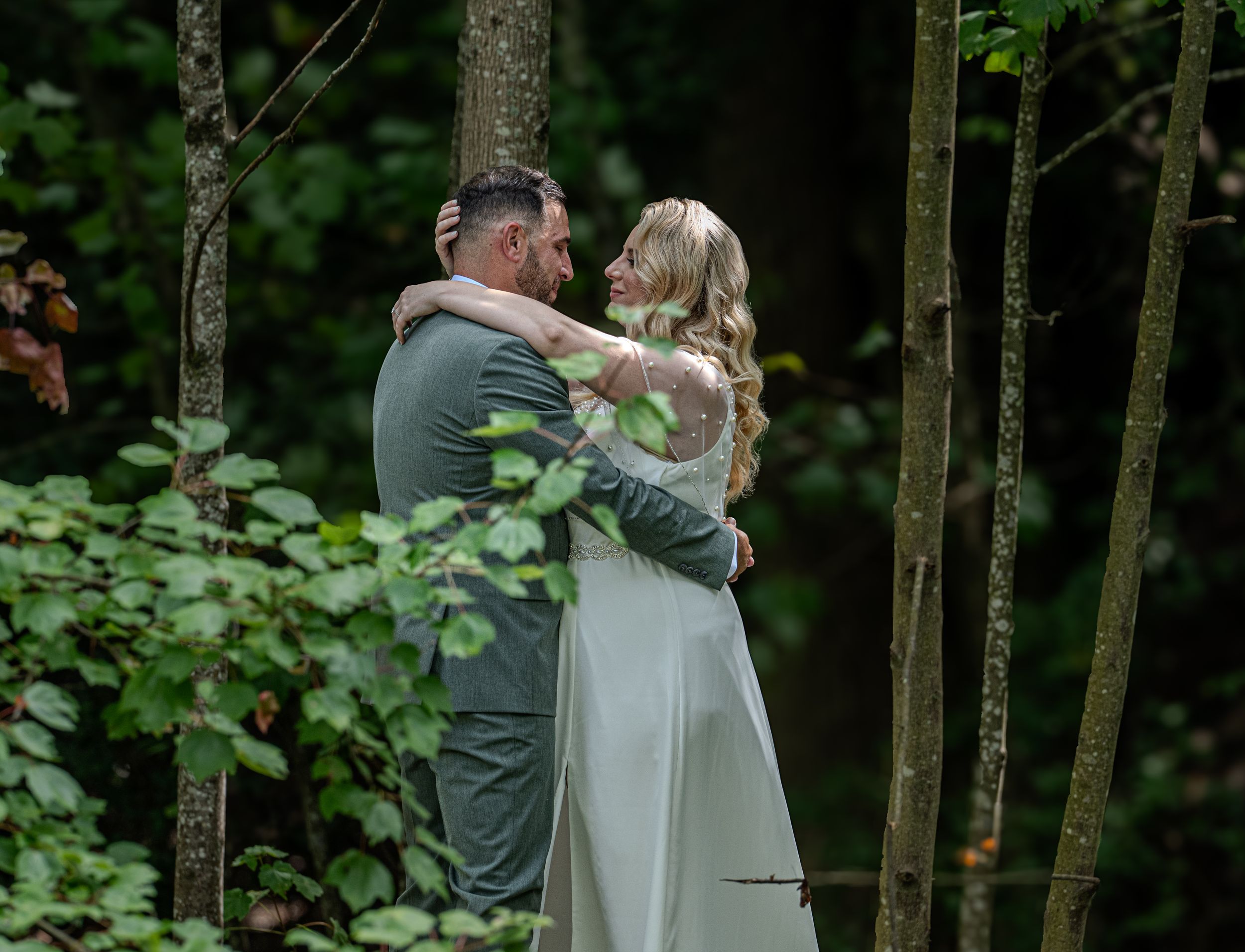 A romantic couple embraces in a serene forest setting, surrounded by lush greenery and tree trunks.