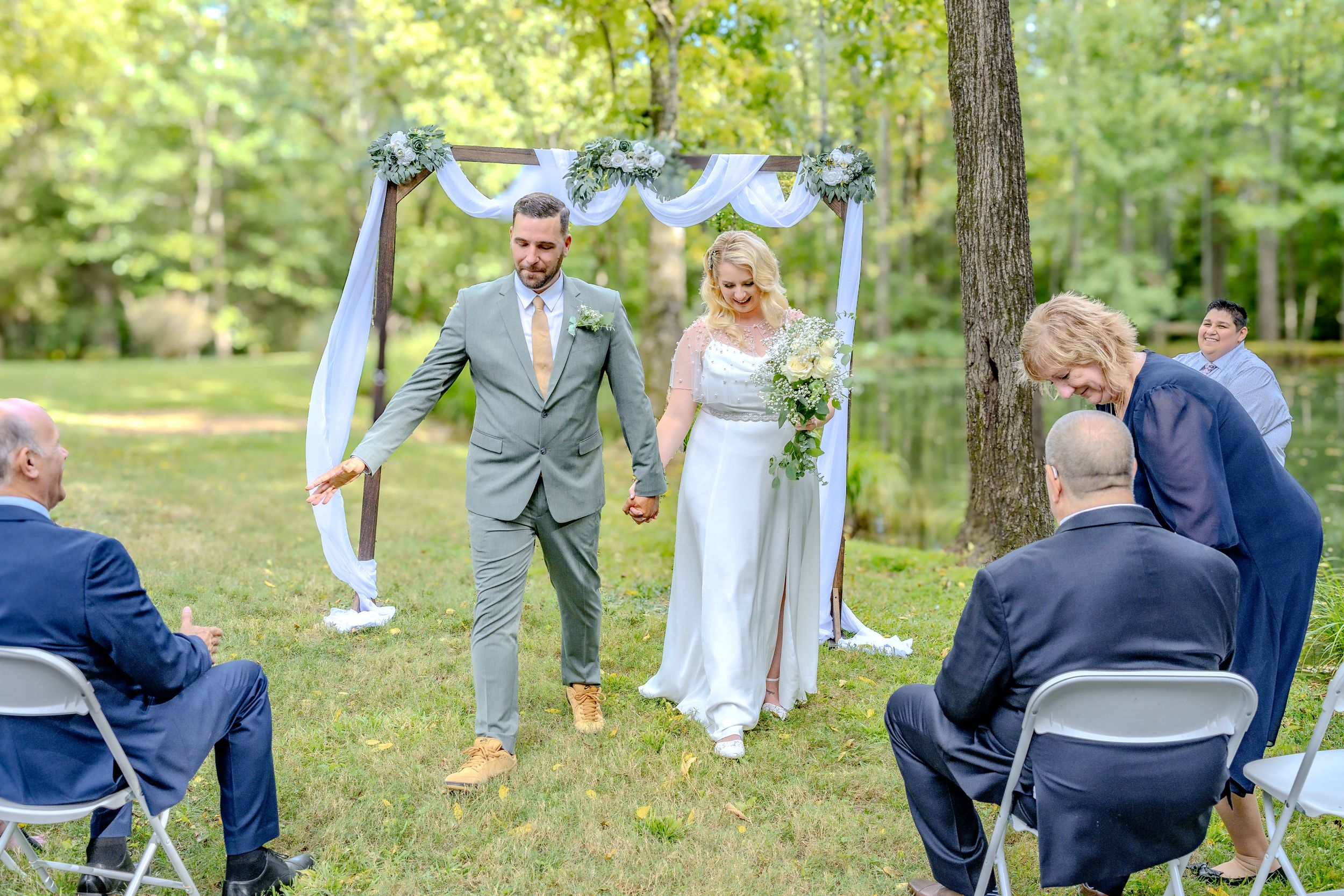 Outdoor wedding ceremony in a forest setting with decorative blue and white arch during a sunny summer day.