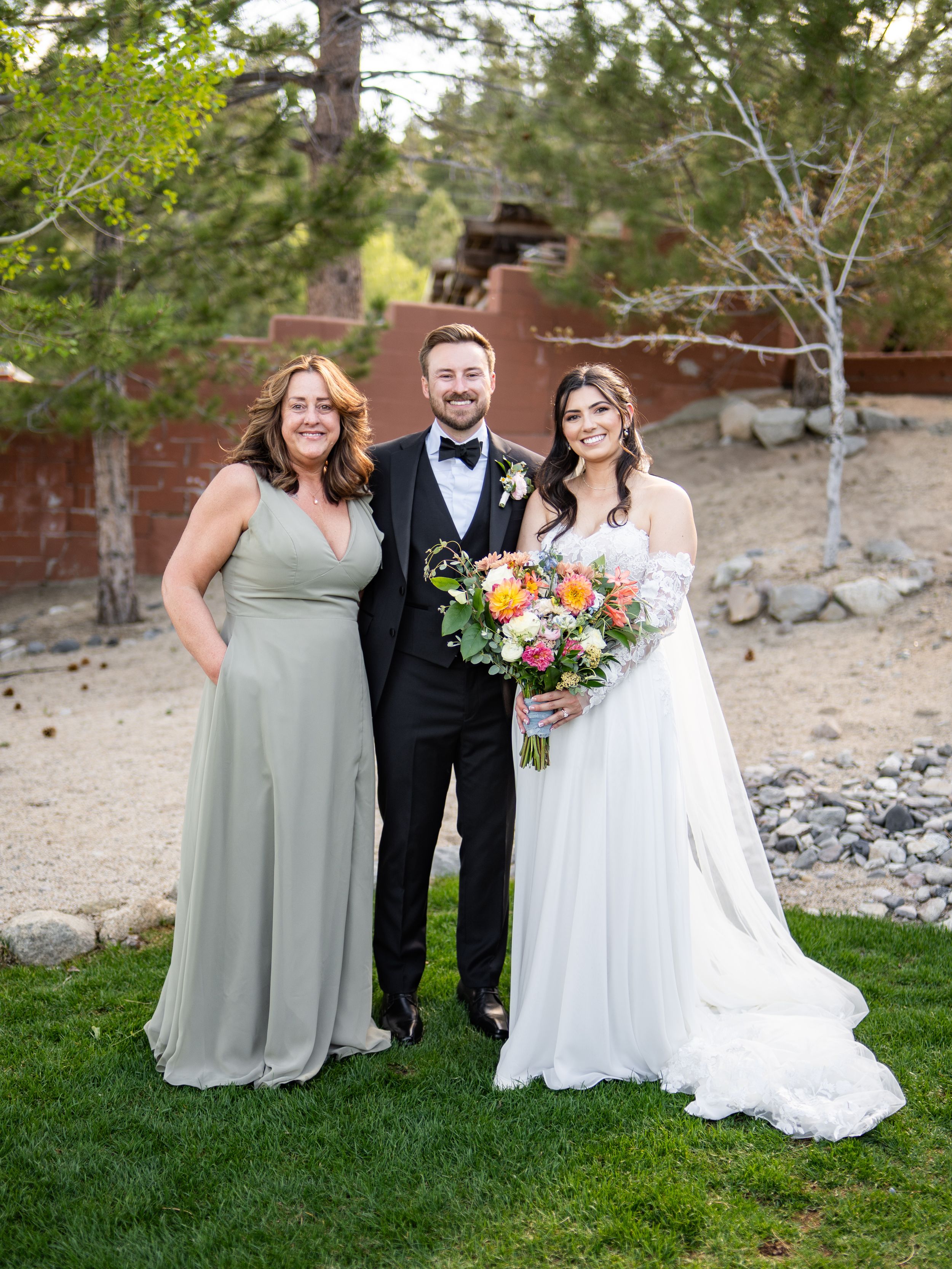 Happy couple poses with wedding party member in sage green dress on grassy outdoor venue with stone wall backdrop.