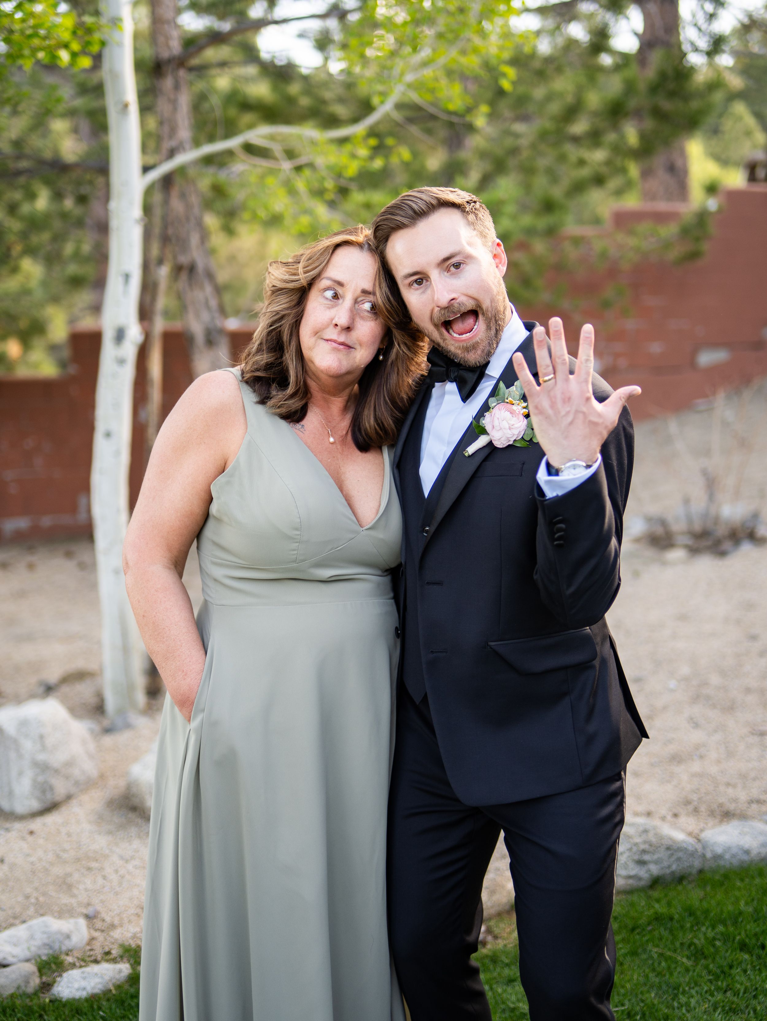 Wedding guests in formal attire pose together outdoors, with a woman in sage green dress and man in black tuxedo.
