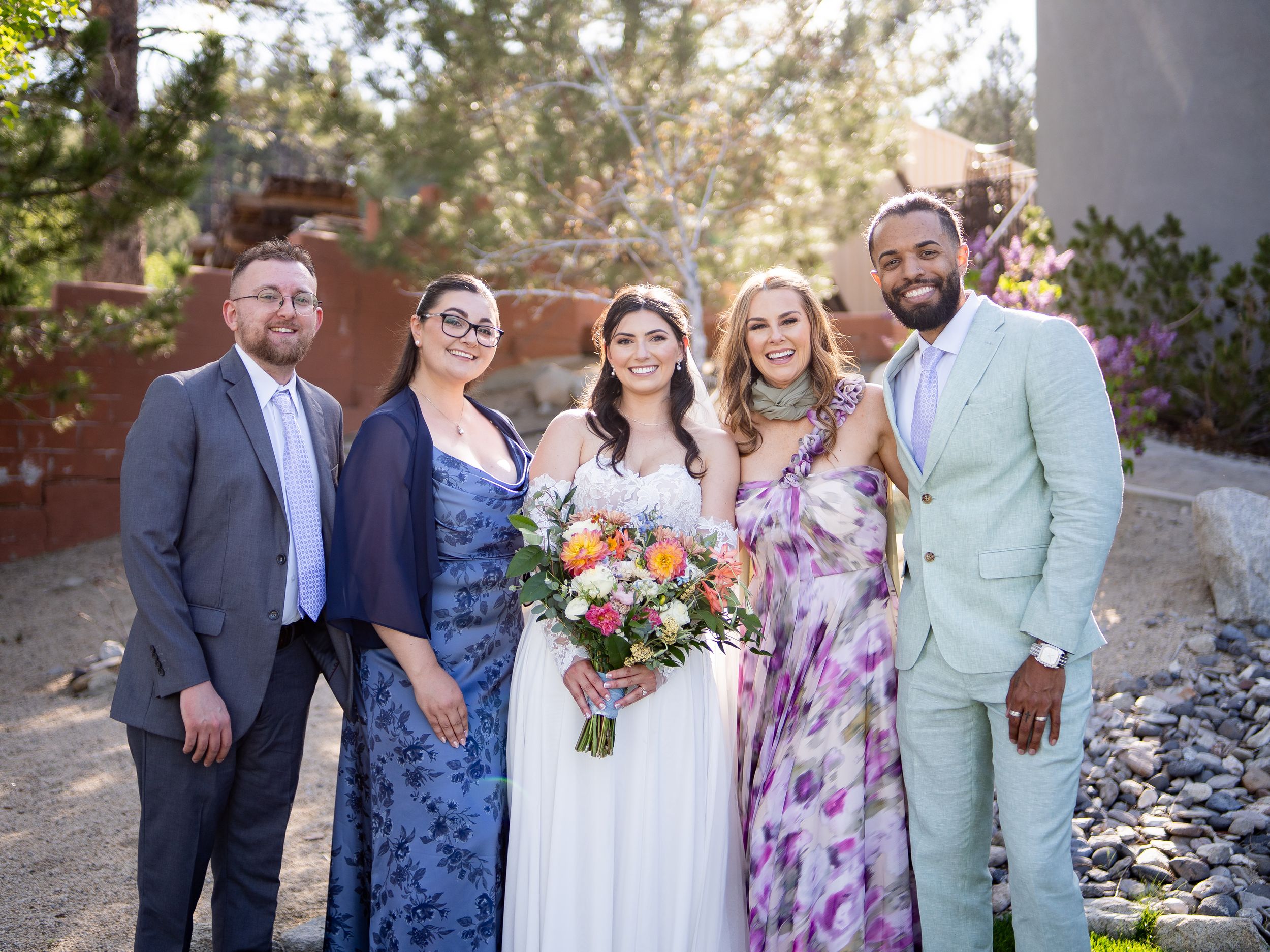 A group stands together outdoors at a wedding celebration, with the bride in white holding a vibrant floral bouquet.