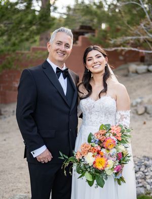 A couple in formal wedding attire stand close together with colorful floral bouquet outdoors at sunset.