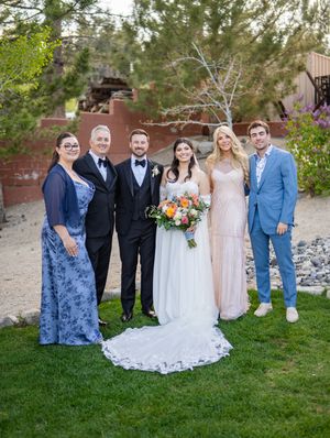 A group of six people in formal attire pose together at an outdoor wedding with a beautiful green backdrop.