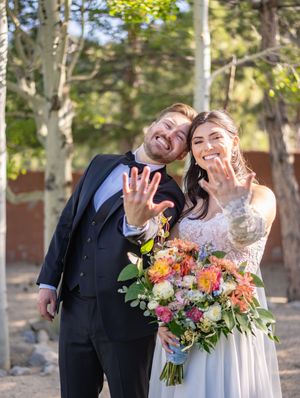 Newlyweds show off their rings while holding a vibrant wildflower bouquet outdoors among aspen trees in natural lighting.