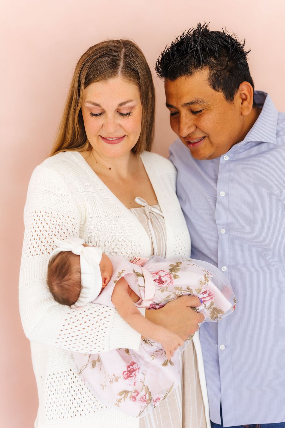 New parents in white and blue attire lovingly gaze at their newborn baby wrapped in a floral blanket against a pink background.