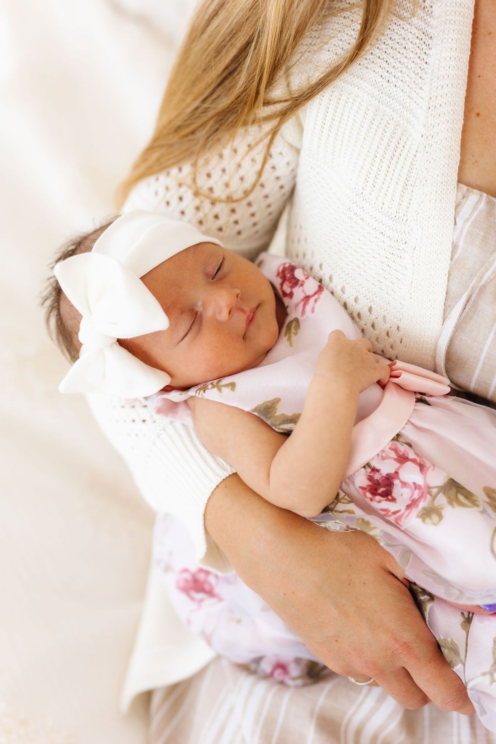A sleeping baby in a white bow and floral dress lies cradled in someone's arms wearing a cream knit sweater. newborn photographer in utah