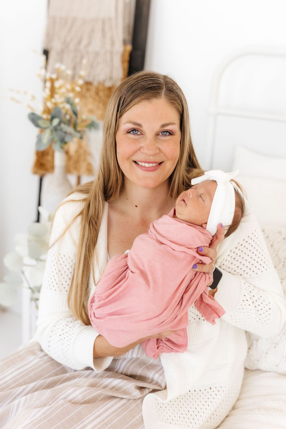 A woman in a white knit sweater holds a sleeping baby wearing pink on a bed with neutral bedding. newborn photographer in utah newborn photo