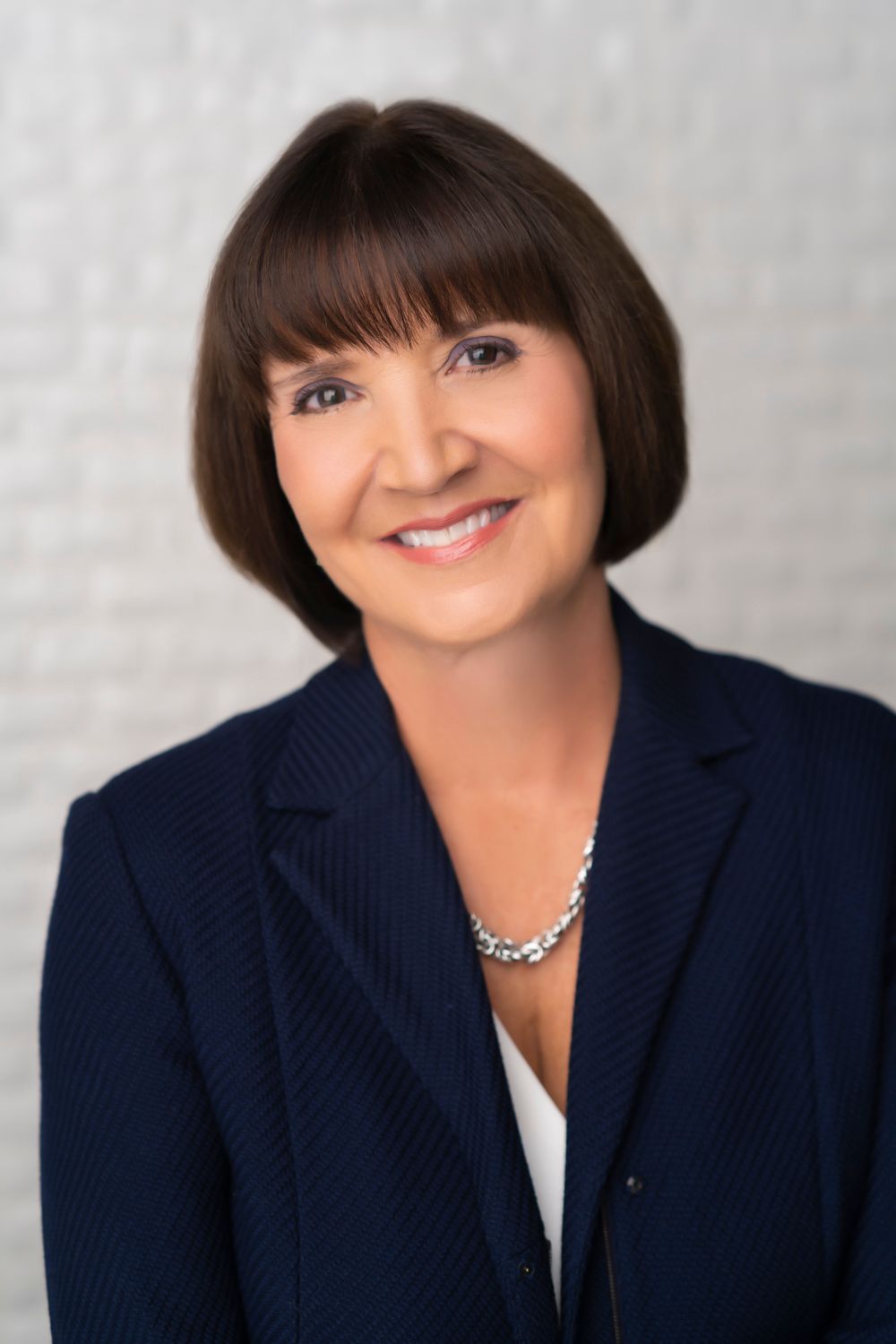Professional headshot of a person in navy blazer with silver necklace against white background.