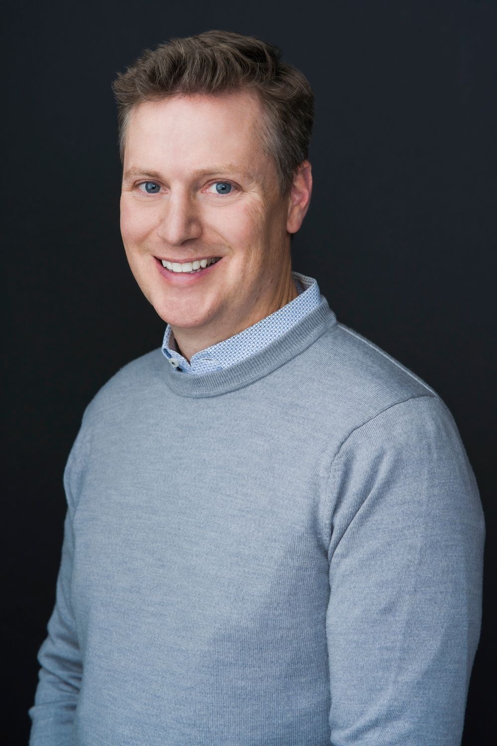 Professional headshot of man in gray sweater smiling against dark background.