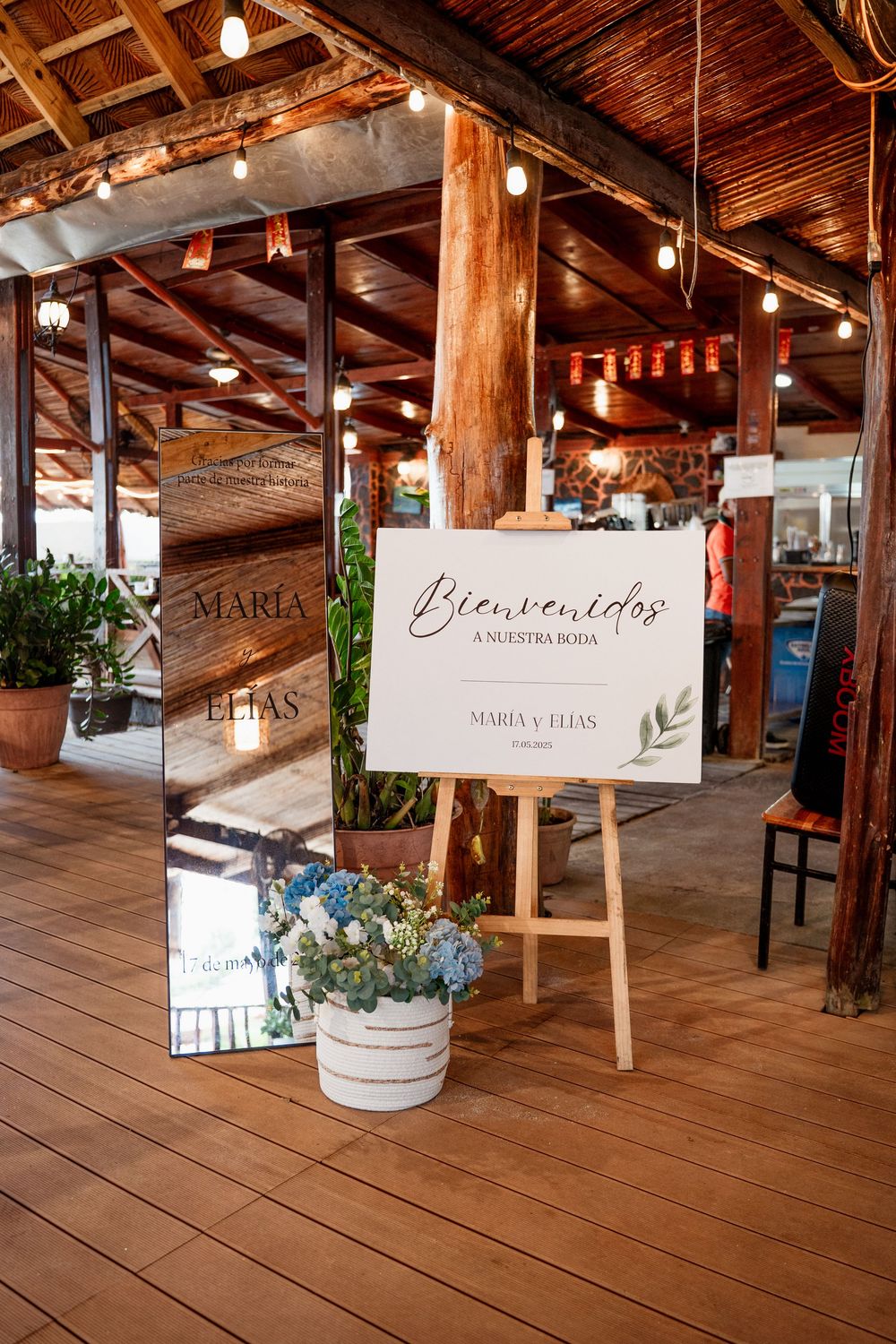 Rustic wooden wedding venue interior with welcome signs on easels and string lights hanging from exposed beam ceiling.
