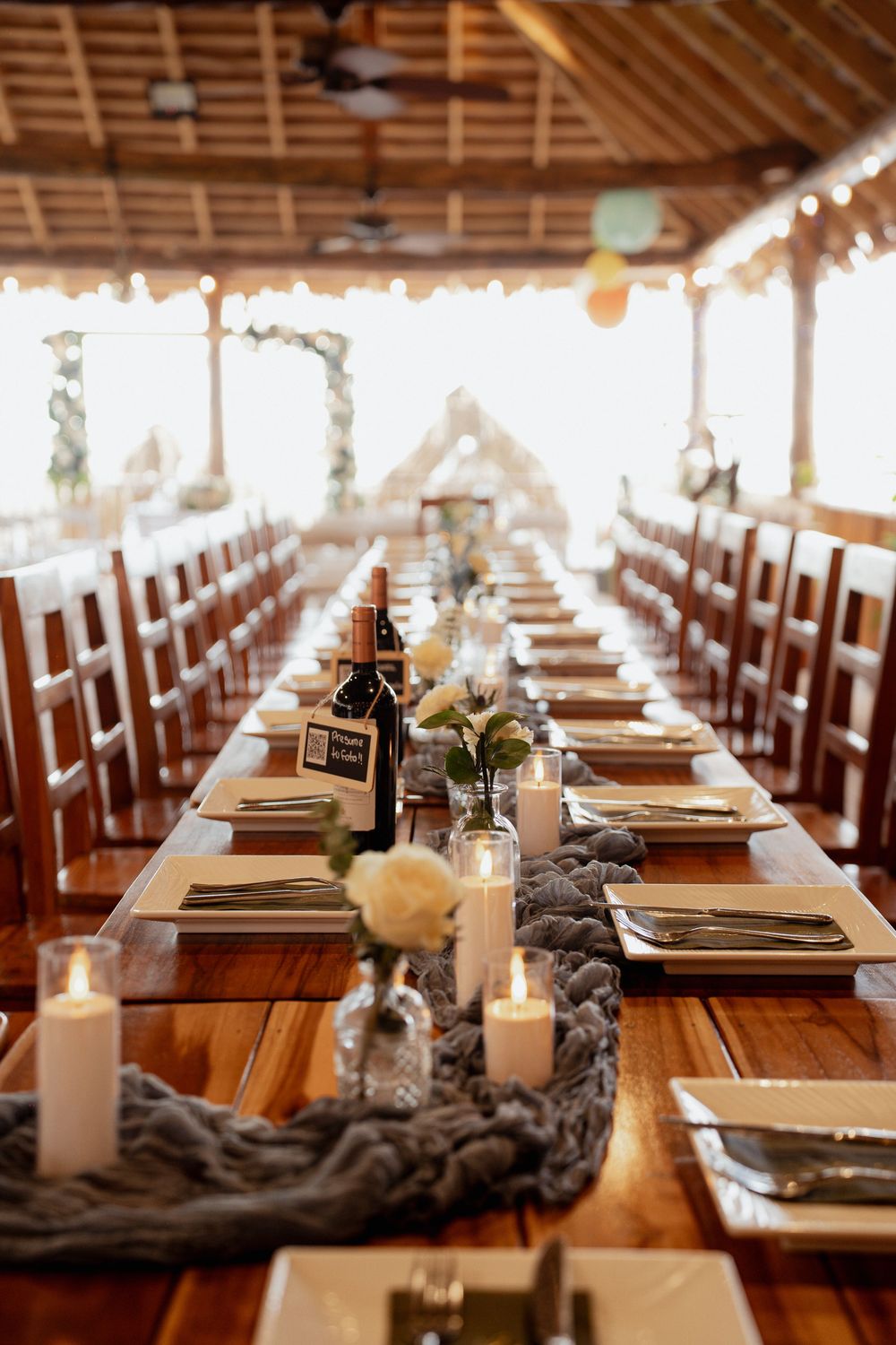 Long wooden reception table set for wedding dinner with candles, gray runner and elegant place settings in rustic covered venue.