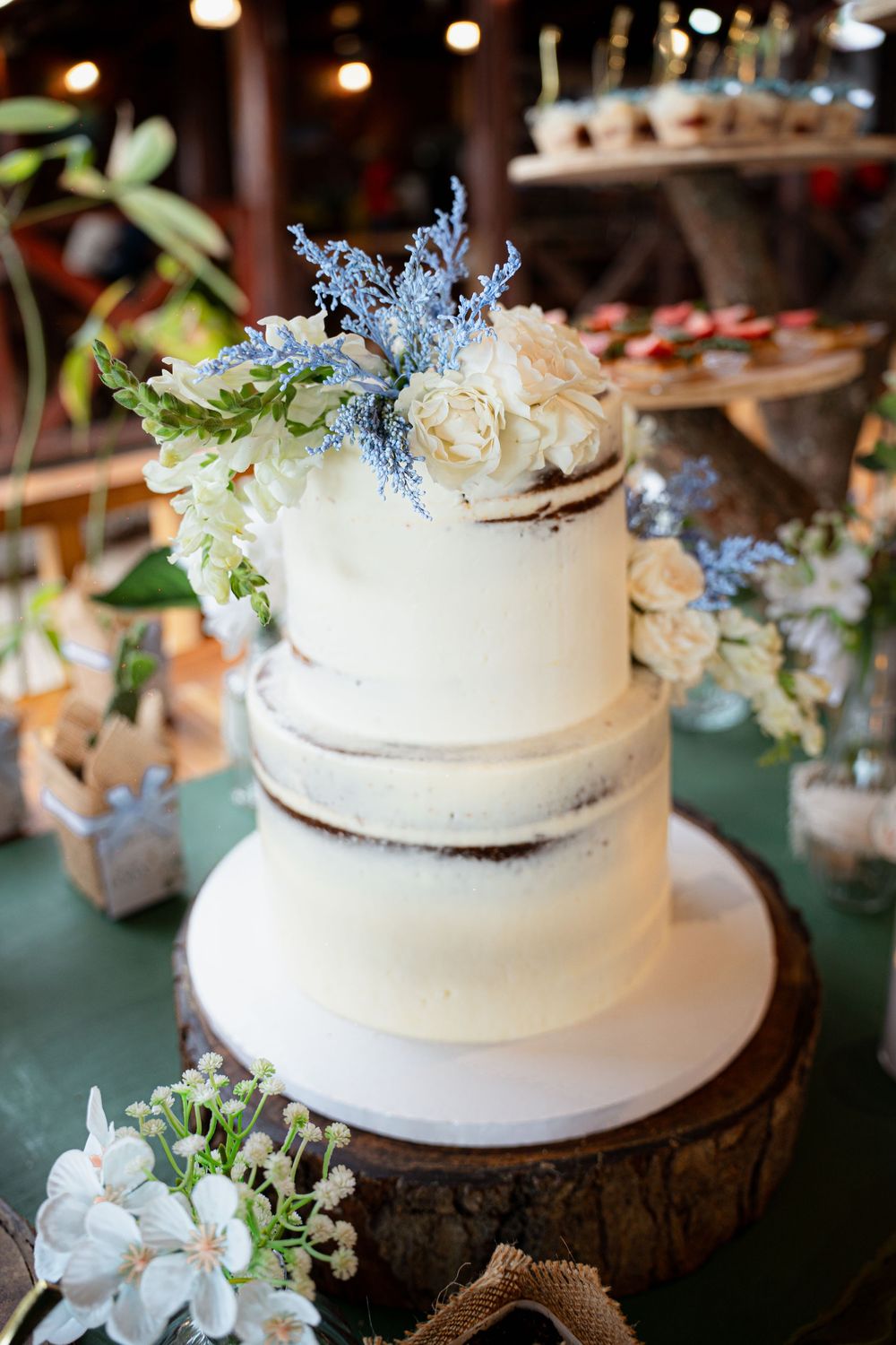 A beautiful white tiered wedding cake decorated with fresh flowers and blue accents sits on a rustic wooden base.