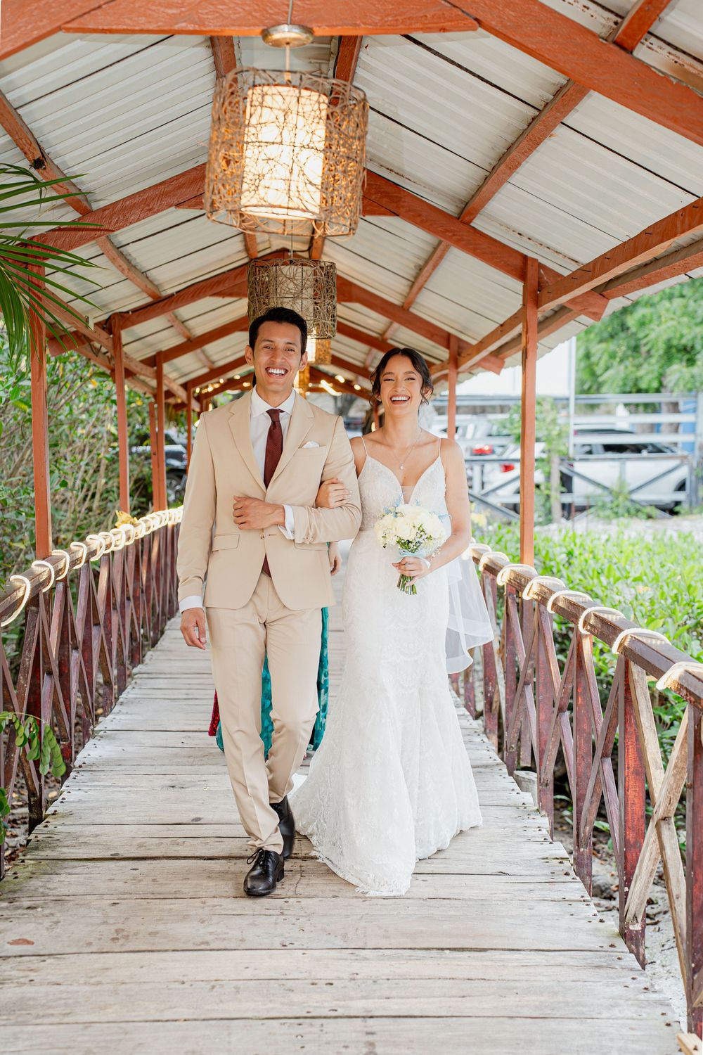 A couple walks down a covered wooden bridge walkway decorated with rustic lanterns during their wedding celebration.