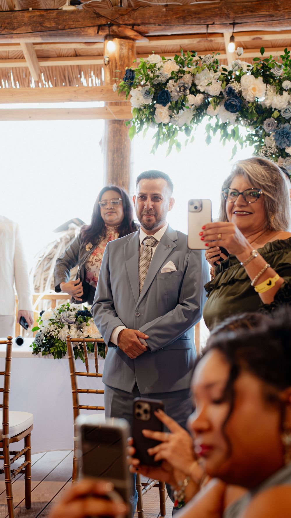 Wedding guests use their phones to capture a special moment during an indoor ceremony with floral decorations.