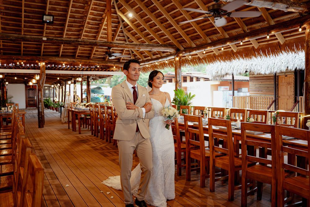 Long wooden tables and benches fill an open-air beach restaurant with thatched roof and tropical decor.
