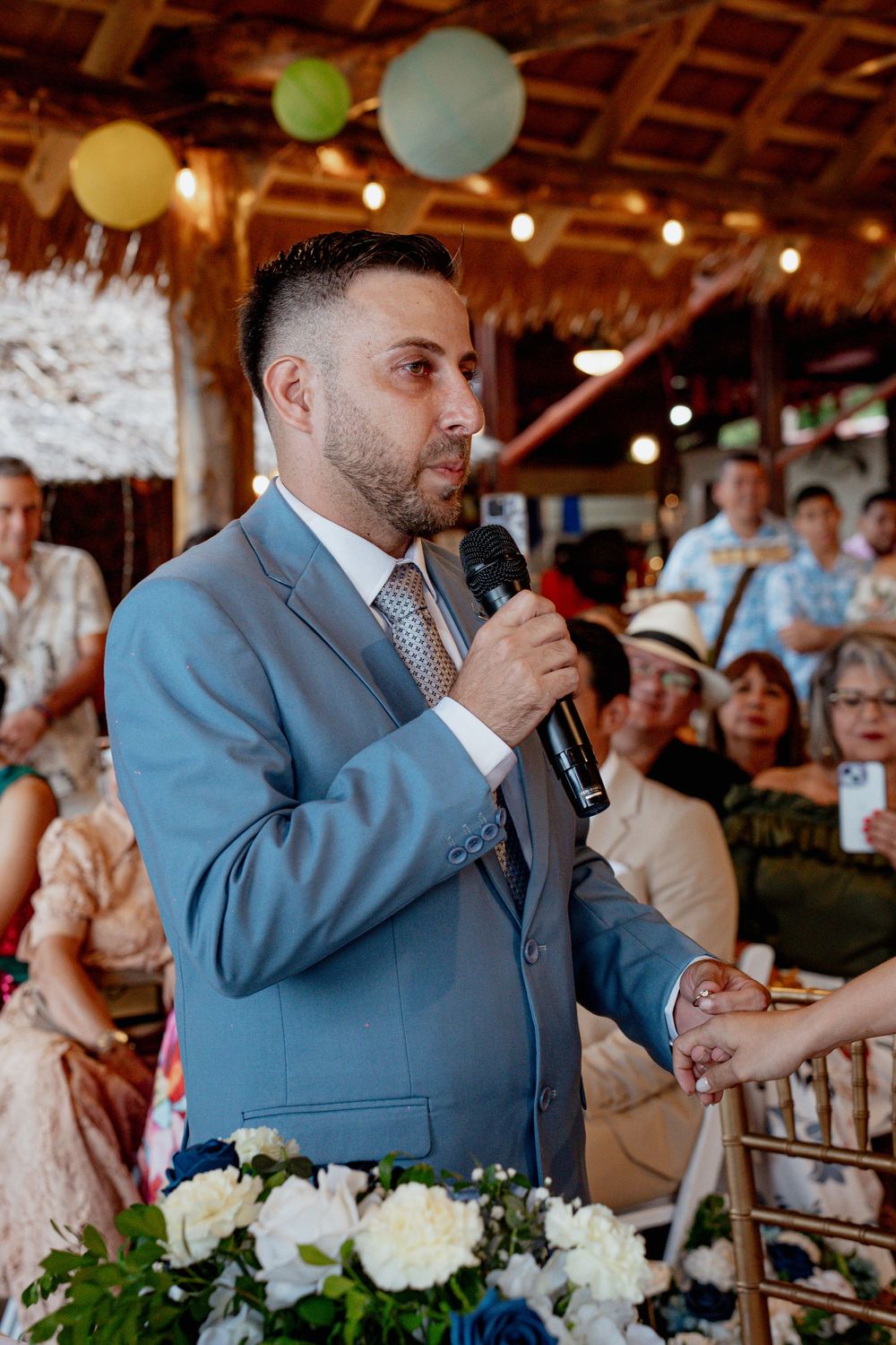 A speaker in a light blue suit gives remarks at a wedding reception decorated with balloons and floral centerpieces.