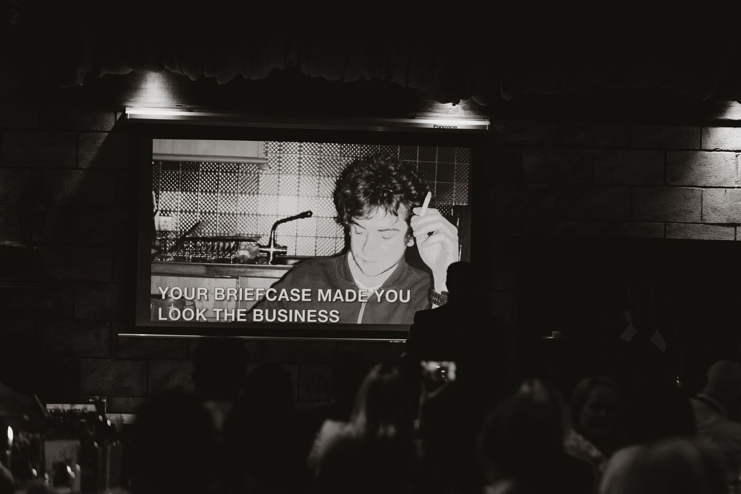 Black and white photo showing a projection screen displaying text in a darkened performance venue or theater space.