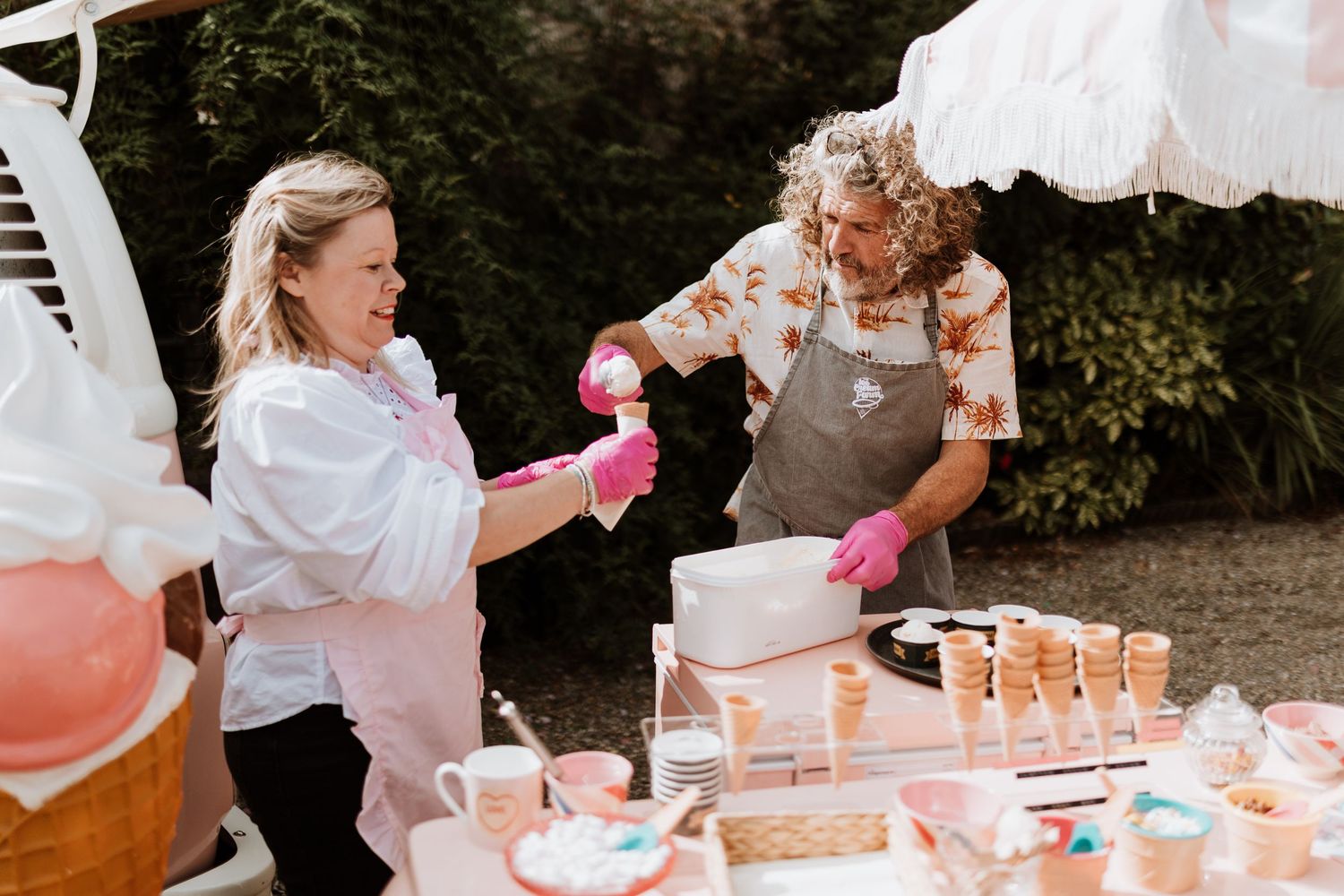 Two people in aprons scoop icecream at an outdoor drink station with cups and pitchers on a pink-themed table.