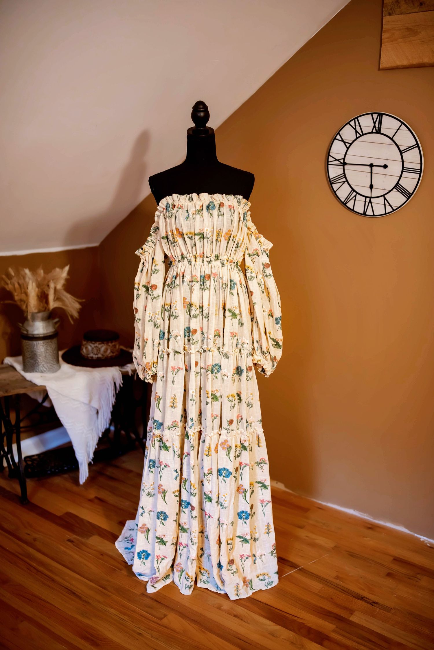 A vintage-style floral off-shoulder dress displayed on a black mannequin with a clock on the tan wall behind it.